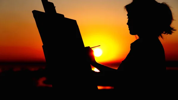 Σιλουέτα μιας γυναίκας που ζωγραφίζει μια εικόνα με βούρτσα μπογιάς και παλέτα σε καμβά σε ένα καβαλέτο, προφίλ κορίτσι πρόσωπο που ασχολούνται με την τέχνη σε ένα χωράφι στο ηλιοβασίλεμα — Φωτογραφία Αρχείου