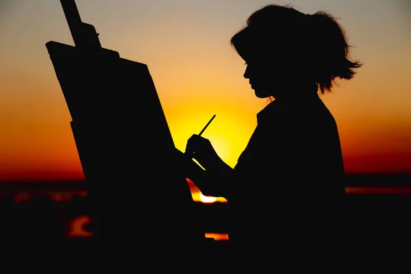 Silhouette einer Frau, die ein Bild auf Leinwand auf einer Staffelei malt, Mädchen mit Pinsel und Palette, die sich bei Sonnenuntergang auf einem Feld mit Kunst beschäftigt — Stockfoto