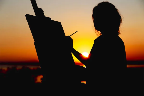 Sziluettje egy nő festmény egy képet a vásznon egy festőállvány, lány ecset és a paletta foglalkozó művészeti területen naplementekor — Stock Fotó