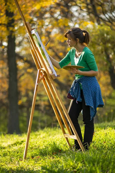 Mujer dibujando un cuadro sobre lienzo sobre un caballete en la naturaleza, una chica con un pincel y una paleta de pinturas en hermoso paisaje, un concepto de pasatiempo — Foto de Stock