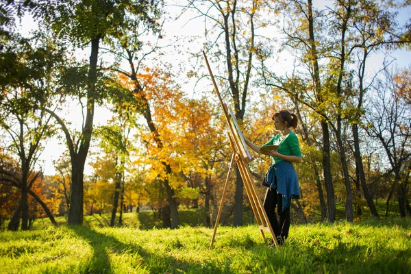 Mujer dibujando un cuadro sobre lienzo sobre un caballete en la naturaleza, una chica con un pincel y una paleta de pinturas en hermoso paisaje, un concepto de pasatiempo — Foto de Stock