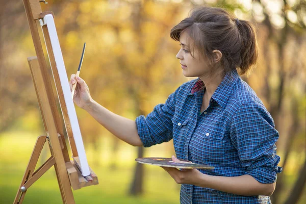 Femme artiste dessiner une image sur un chevalet dans la nature, une fille avec un pinceau et une palette, un concept de créativité et un passe-temps — Photo