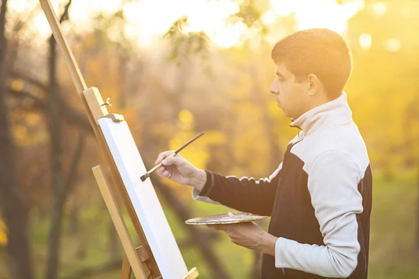 Young artist paints a picture on canvas on an easel in nature, a man with a brush and a a palette of paints among autumn trees, — 스톡 사진
