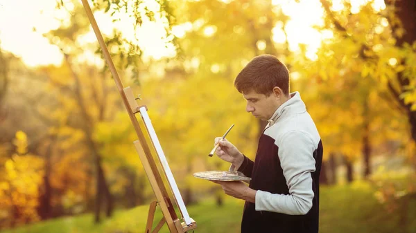 Fiatal művész rajzol egy képet vászonra egy festőállványra a természetben, egy férfi ecsettel és egy festő gamuttal az őszi fák között, a szabadidős tevékenység fogalma — Stock Fotó