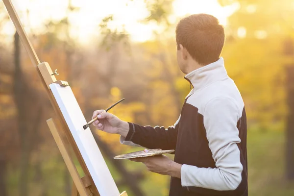 Genç ressam tuvale bir resim çizer doğadaki bir sehpaya, fırçalı ve palet boyalı bir adam sonbahar ağaçları arasında., — Stok fotoğraf