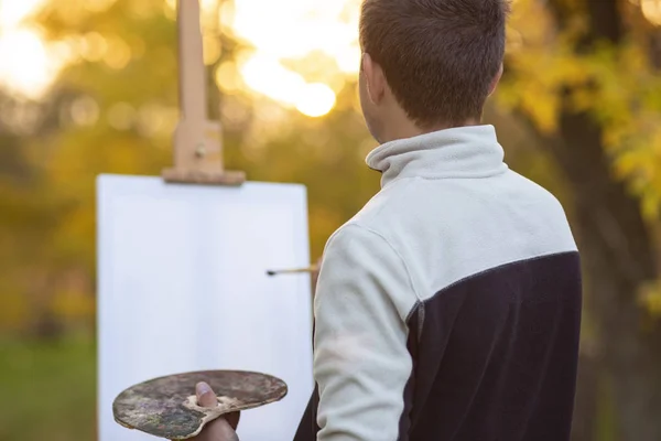 Joven artista pinta un cuadro sobre lienzo sobre un caballete en la naturaleza, un hombre con un pincel y una paleta de pinturas entre los árboles de otoño , — Foto de Stock