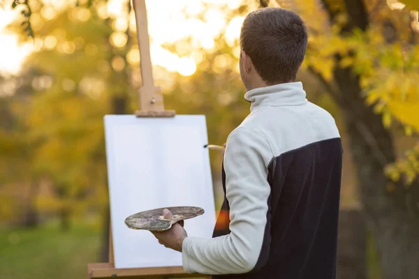 Fiatal művész festett egy képet vászonra egy festőállványra a természetben, egy férfi ecsettel és egy festékpalettával az őszi fák között, — Stock Fotó