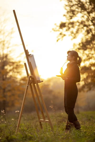 Semi silueta de la joven artista pintando un cuadro en el caballete, una figura con un pincel y una paleta en el fondo del paisaje otoñal — Foto de Stock