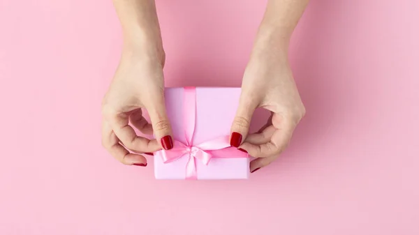 Menina segurando um presente nas mãos, as mãos das mulheres abrem a caixa envolta em papel decorativo em um fundo rosa pastel, vista superior, férias conceituais e presentes — Fotografia de Stock