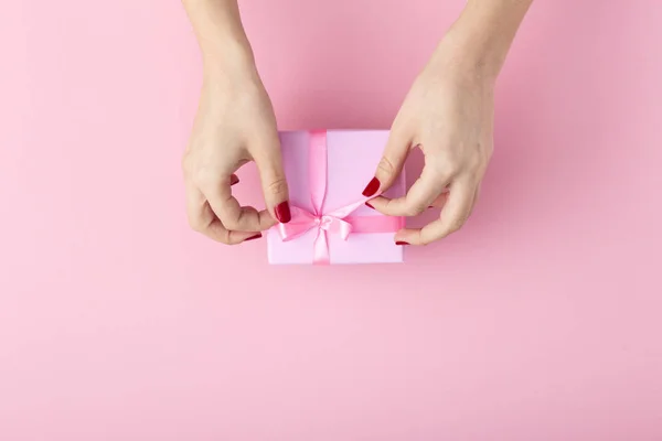 Menina segurando um presente nas mãos, as mãos das mulheres abrem a caixa envolta em papel decorativo em um fundo rosa pastel, vista superior, férias conceituais e presentes — Fotografia de Stock