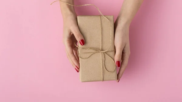 Mãos femininas com uma caixa de presente em uma vista superior de fundo colorida pastel, a menina segurando um presente envolto em papel artesanal decorativo, o conceito de feriados — Fotografia de Stock