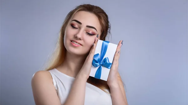 Portrait of a happy woman with a gift box near the face on a studio background, the girl closed her eyes from pleasure and pressed a present with bow to her cheek, concept holidays and love and care — Stock Photo, Image