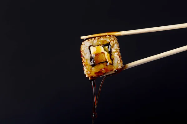 succulent roll between chopsticks on black background, drops of soy sauce dripping from sushi, food background, Japanese cuisine