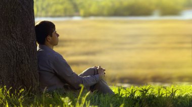 Genç adam sırtı ağaca dönük oturuyor ve hayat, din ve dinlenme kavramı hakkında meditasyon yapıyor.