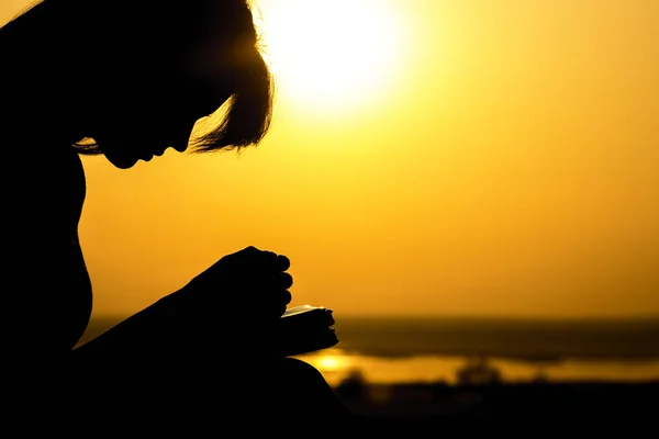 Silhueta das mãos da mulher orando a Deus na natureza witth a Bíblia ao pôr do sol, o conceito de religião e espiritualidade — Fotografia de Stock