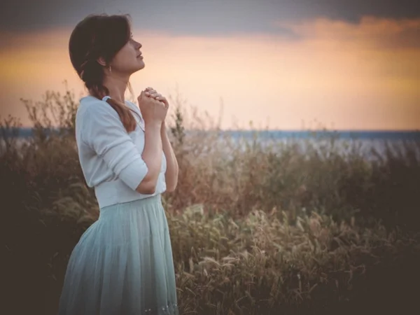 Профиль силуэта красивой девушки в платье, молящейся Богу в поле, молодая женщина, идущая по природе во время заката глядя в небо, религиозная концепция — стоковое фото