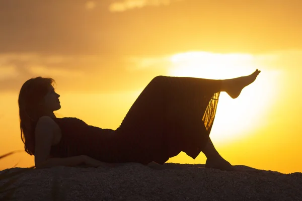 Silueta de una joven hermosa chica acostada en un vestido en la arena y disfrutando de la puesta de sol, la figura de una mujer en la playa, el concepto de vacaciones de verano, el turismo , —  Fotos de Stock