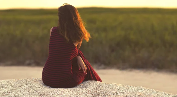 Joven chica de ensueño sentado en la arena y mirando a la distancia en el campo en el horizonte, una mujer disfrutando de la puesta de sol y la naturaleza, el concepto de salud y relajación —  Fotos de Stock