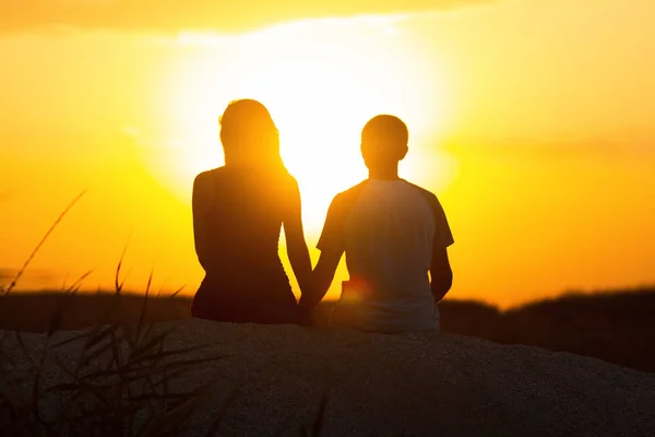 Silhouette of a loving couple at sunset sitting on sand on the beach, the figure of a man and a woman holding hands, in love, a romantic scene in nature, summer rest — Stock Photo, Image