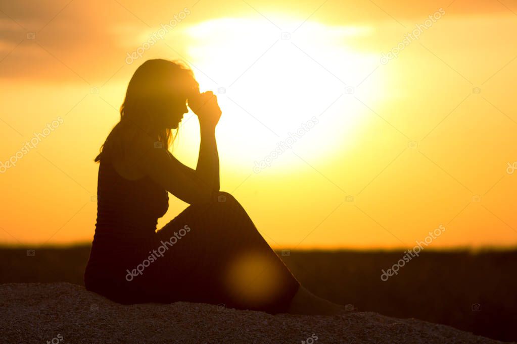 silhouette of girl sitting on the sand and praying to God at sunset, the figure of young woman on the beach, concept religion