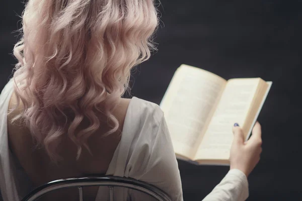 Blonde meisje houdt open boek zittend op een stoel, terug vanjonge vrouw het lezen van een roman, concept van hobby, ontspanning, vrije tijd op zwarte achtergrond — Stockfoto