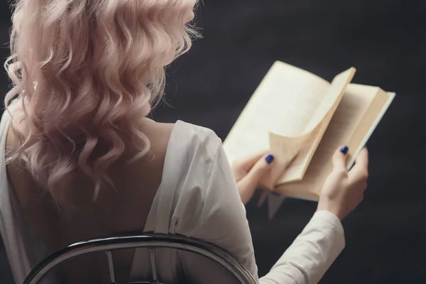 Blonde meisje houdt open boek zittend op een stoel, terug vanjonge vrouw het lezen van een roman, concept van hobby, ontspanning, vrije tijd op zwarte achtergrond — Stockfoto