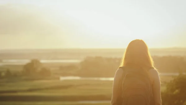 Giovane ragazza con uno zaino sulle spalle seduta in un campo e godersi il tramonto, concetto di escursioni, passeggiate, riposo, stile di vita attivo — Foto Stock