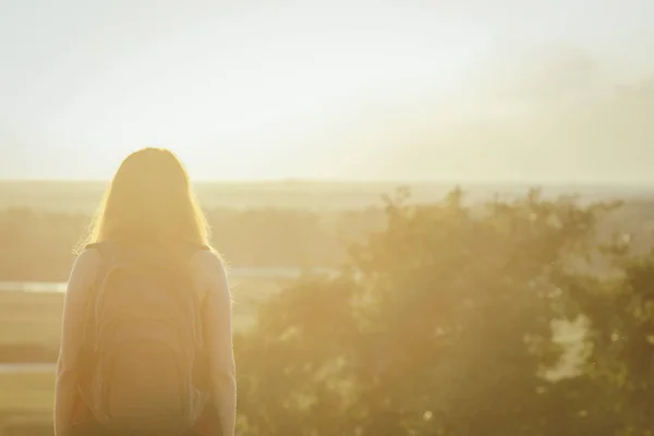 Молодая девушка с рюкзаком на плечах сидит в поле и наслаждается закатом, концепция походов, ходьба, отдых, активный образ жизни — стоковое фото