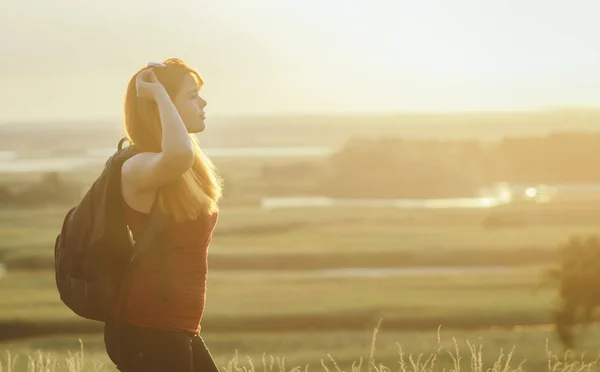 Молодая девушка с рюкзаком на плечах сидит в поле и наслаждается закатом, концепция походов, ходьба, отдых, активный образ жизни — стоковое фото