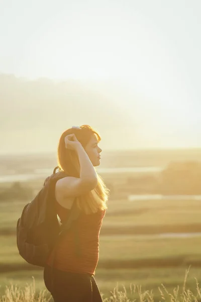 young girl with a backpack on her shoulders sitting in a field and enjoying the sunset, concept of hiking, walking, rest, active lifestyle