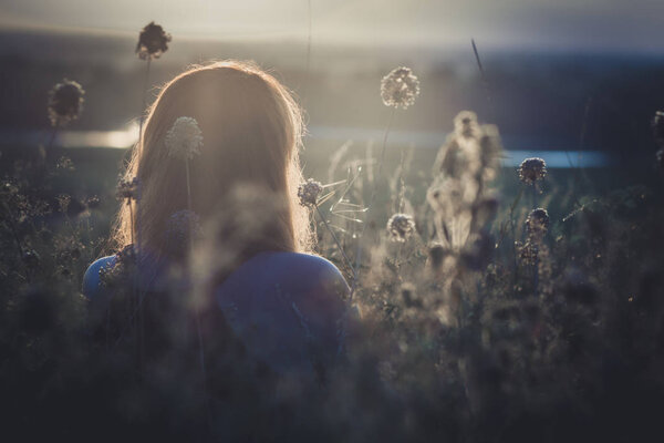 Head backof red-haired girl sitting on the ground in a field among dried flowers and enjoys nature at sunset, young woman relaxing, concept of rest, healthcare, harmony, lifestyle
