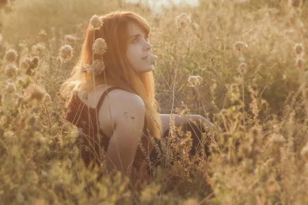 Dromerig roodharige meisje zit op de grond in een veld onder gedroogde bloemen en geniet van de natuur bij zonsondergang, jonge vrouw ontspannen, concept van rust, gezondheidszorg, harmonie, levensstijl — Stockfoto