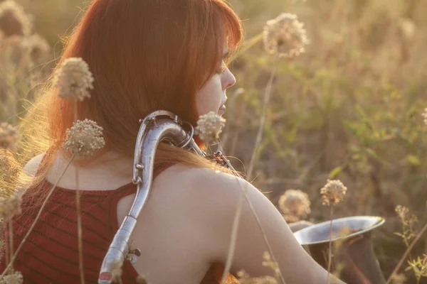 Dreamy red-haired girl with saxophone on the shoulder sitting on the ground in a field among dried flowers and enjoys nature at sunset, young woman relaxing, concept of relax, lifestyle — Stock Photo, Image