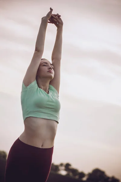 young woman in sportswear exercises and stretches hands up on background of cloudy sky, girl engaged in sport outdoors, concept healthy lifestyle and bodycare