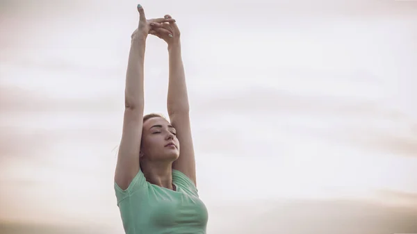 young woman in sportswear exercises and stretches hands up on background of cloudy sky, girl engaged in sport outdoors, concept healthy lifestyle and bodycare