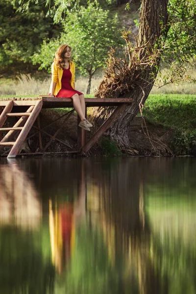Escena romántica de mujer joven sentada en un puente de madera del río con un libro cercano y disfrutando de la naturaleza otoñal, chica relajante caminando al aire libre — Foto de Stock