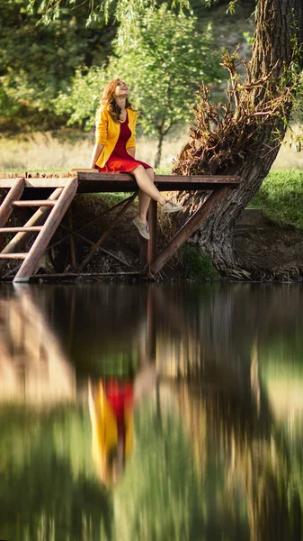 Porträt eines glücklichen und unbeschwerten Mädchens, das am Flussufer auf einer Holzplattform sitzt, die Beine baumeln lässt und lacht, junge Frau mit einem Buch, das die Natur genießt, die sich im Wasser spiegelt — Stockfoto