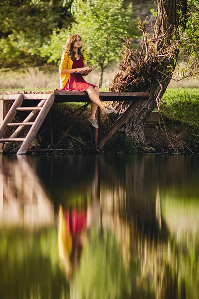 Hermosa joven en las orillas del río sentado en una plataforma de madera con un libro, adolescente leyendo literatura de ficción sobre la naturaleza, hobby concepto — Foto de Stock