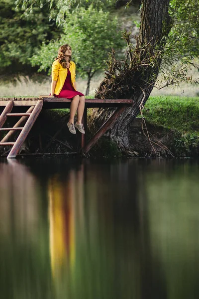 Escena romántica de una joven mujer pensativa sentada en un puente de madera con un libro cerca y mirando hacia arriba, reflejo femenino en el agua — Foto de Stock