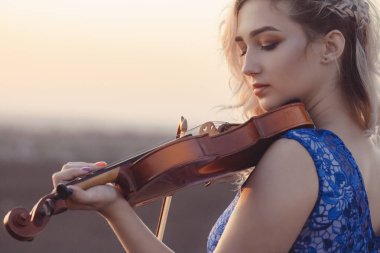 Gün batımında çenesinin altında kemanla güzel bir genç kadının portresi, doğada müzik aleti çalan bir kız, konsept müzik ve hobiler.