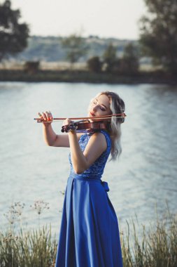 Şafakta nehir kıyısında kemanı olan romantik genç bir kadın, uzun elbiseli zarif bir kız müzik sanatıyla meşgul, doğa üzerine performans.
