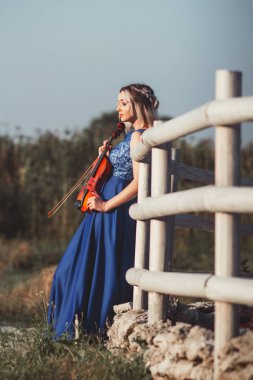 Uzun elbiseli güzel bir kızın portresi ahşap bir çitin yanında bir kemanla, genç bir kadın açık havada müzik aleti ile yürüyor.