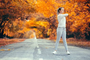 Spor kıyafetleri ve asfalt yolda ısınma egzersizleri yapan sıska genç bir kadın sonbahar doğası, konsept sağlık ve aktif yaşam tarzı arka planında sporla meşgul olan bir kız.