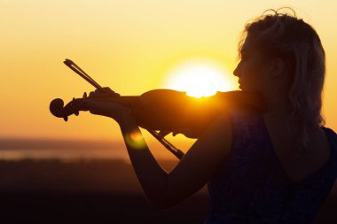 Gün batımında keman çalan bir kadın figürünün yüz profili silueti, doğa performansı, çenesinin altında müzik aleti olan bir kız, konsept müzik sanatı
