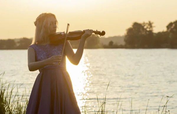 Silueta romántica de mujer joven con un violín al amanecer en la orilla del río, chica elegante tocando un instrumento musical en la naturaleza, concepto de música y la inspiración — Foto de Stock