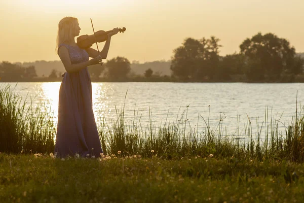 Silueta romántica de mujer joven con un violín al amanecer en la orilla del río, chica elegante tocando un instrumento musical en la naturaleza, concepto de música y la inspiración — Foto de Stock