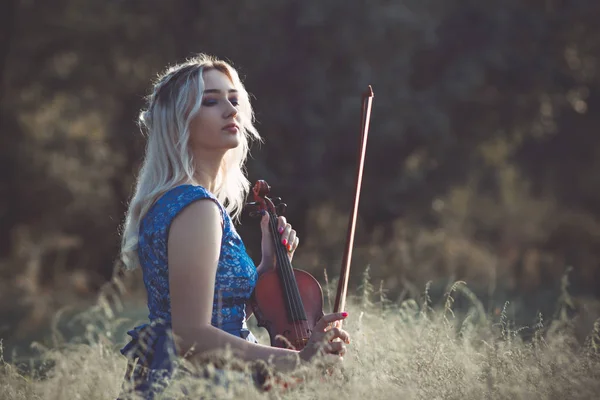 새벽에 마른 잔디에 앉아 있는 바이올린을 든 드레스를 입고 멋지고 아름다운 소녀의 초상화, 자연에서 편하게 하는 영감을 주는 악기를 연주하는 여성 — 스톡 사진