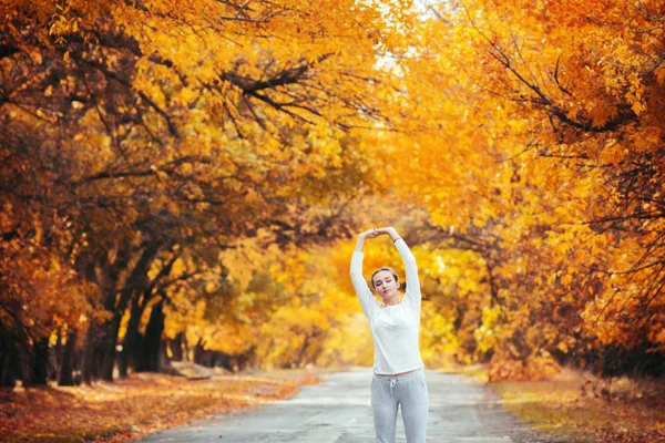 スポーツウェアの練習でスリムな若い女性とアスファルトの道路に立って手を伸ばし 女の子は秋の葉を背景に屋外スポーツに従事し コンセプト健康的なライフスタイルと女性の美しさ — ストック写真