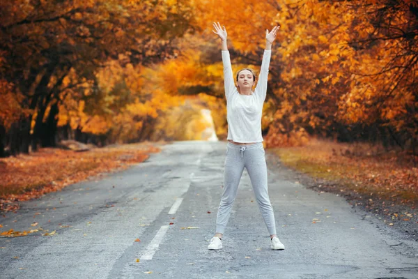 スポーツウェアの練習でスリムな若い女性とアスファルトの道路に立って手を伸ばし 女の子は秋の葉を背景に屋外スポーツに従事し コンセプト健康的なライフスタイルと女性の美しさ — ストック写真