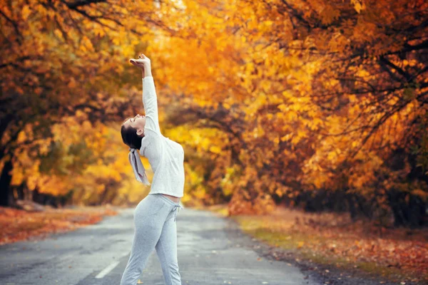 Szczupła młoda kobieta w ćwiczeniach odzieży sportowej i wyciąga ręce do góry satanding na asfaltowej drodze, dziewczyna zaangażowana w sport na świeżym powietrzu na tle jesiennych liści, koncepcja zdrowego stylu życia i kobiecego piękna — Zdjęcie stockowe